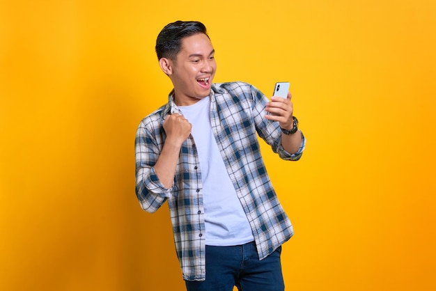 Un joven asiático muy contento con un pantalones a cuadros celebrando el éxito con un teléfono móvil levantando las manos se regocijó de una gran suerte aislado en un fondo amarillo