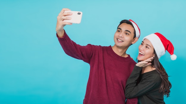 Joven, asiático, pareja, amante, llevando, santa claus, hat., Mano, tenencia, smartphone, selfie, juntos, aislado, en, fondo azul, en, estudio, shot., Navidad, celebración, de, amante, concept.