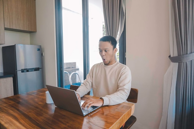Un joven asiático pasa su tiempo en casa sentado en el comedor conmocionado mientras mira su computadora portátil