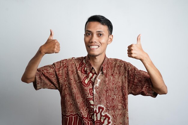 Joven asiático de piel morena con una expresión feliz de camisa batik con alegría y mostrando un pulgar hacia arriba si está de acuerdo con un buen fondo blanco de primera categoría