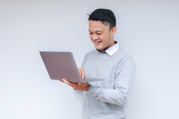 Joven asiático que se siente feliz y sonríe cuando está de pie y trabaja con una computadora portátil Hombre indonesio con camisa gris