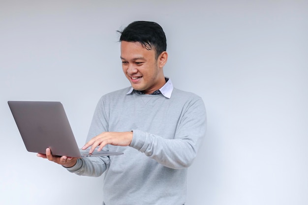 Joven asiático que se siente feliz y sonríe cuando está de pie y trabaja con una computadora portátil Hombre indonesio con camisa gris