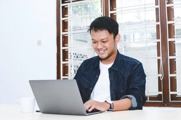 Joven asiático que se siente feliz y sonríe cuando trabaja con una laptop en la mesa Hombre indonesio con camisa azul