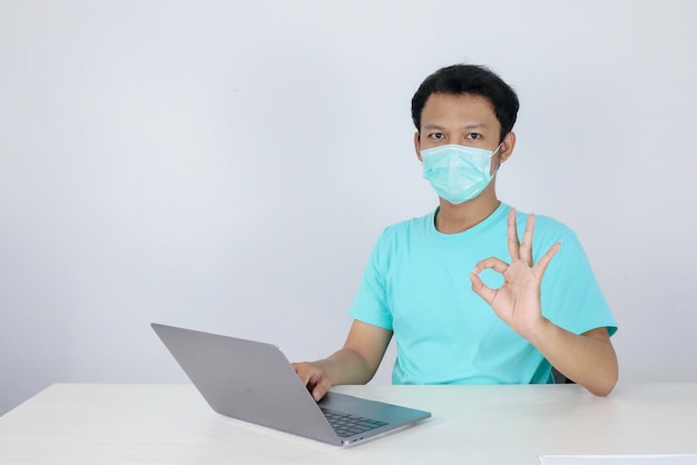 El joven asiático que usa máscara médica es serio y se enfoca cuando trabaja en una computadora portátil en la mesa Hombre indonesio con camisa azul
