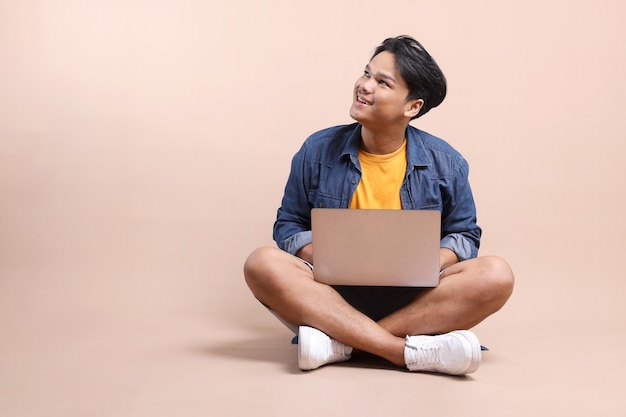 Joven asiático con ropa casual sentado en el suelo con la computadora portátil en sus regazos mirando hacia el emp