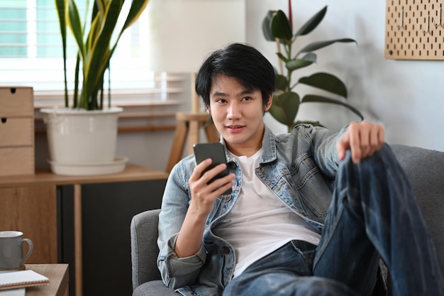 Joven asiático con ropa informal tirado en el sofá y usando el teléfono móvil