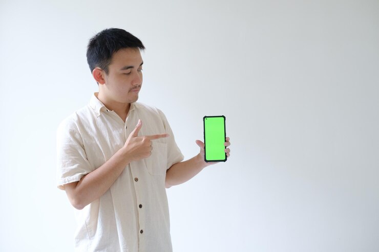 El joven asiático señala y mira un teléfono inteligente con pantalla verde que se sostiene con la mano izquierda