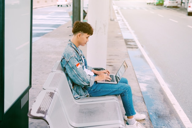 Joven asiático sentado en la silla en la parada de autobús del aeropuerto y usando la computadora portátil, vista lateral