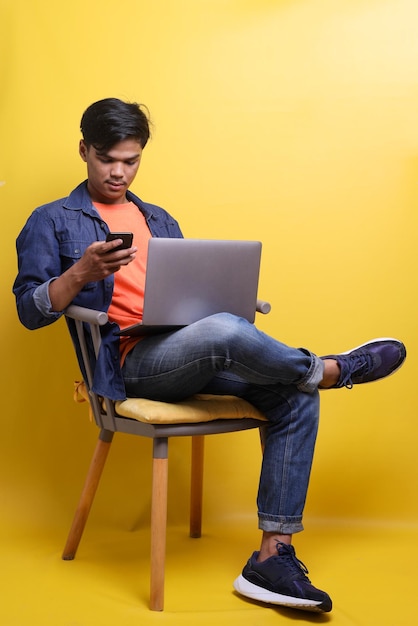 Joven asiático sentado en un sillón usando un teléfono inteligente y una computadora portátil con expresión seria