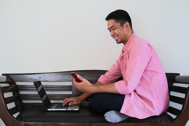 Joven asiático sentado en el sofá de madera mientras trabaja con su computadora portátil y teléfono móvil