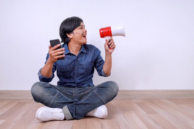 Joven asiático sentado en el suelo gritando a través de un megáfono mientras sostiene un teléfono inteligente