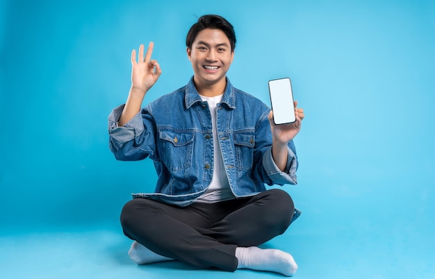 Joven asiático sentado y usando el teléfono con un fondo azul