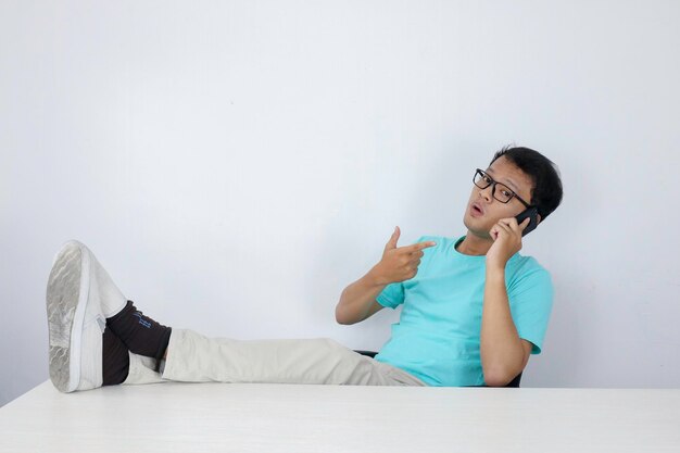 El joven asiático se siente feliz y sonríe cuando habla por teléfono con la pierna sobre la mesa