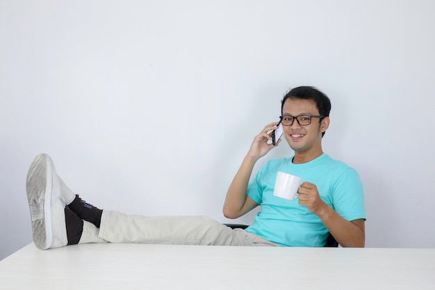 El joven asiático se siente feliz y sonríe cuando habla por teléfono con la pierna sobre la mesa