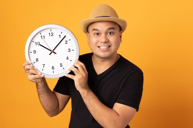 Joven asiático con sombrero, camisa negra sosteniendo un gran reloj de pie sobre un fondo amarillo aislado.