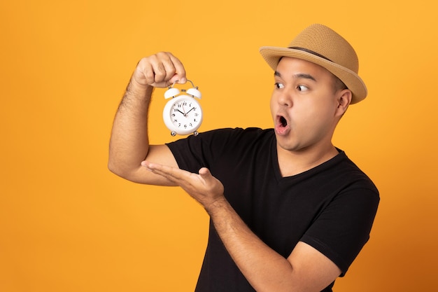 Un joven asiático con un sombrero negro que sostiene un pequeño reloj despertador sobre un fondo amarillo aislado. Se siente feliz y emocionado.