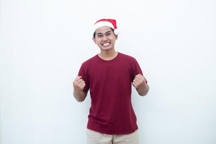 Joven asiático con un sombrero de Papá Noel agarrando ambas manos con entusiasmo y ira