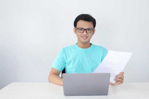 El joven asiático sonríe y es feliz cuando trabaja en una computadora portátil y documenta a un hombre indonesio con camisa azul