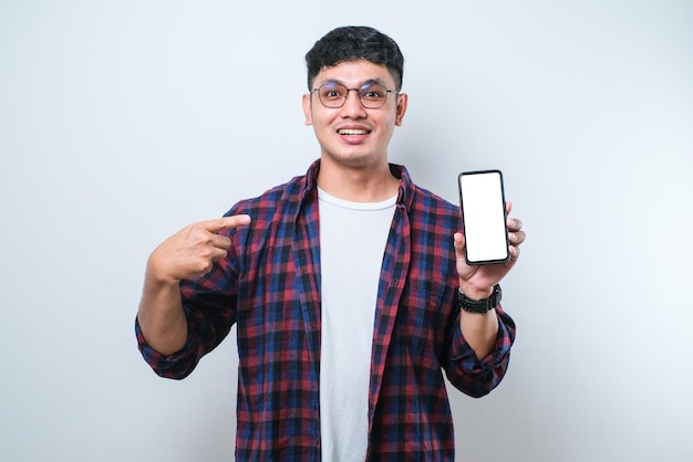 Joven asiático sonríe mirando a la cámara que muestra la pantalla del teléfono inteligente en blanco recomendando la aplicación