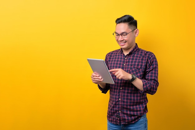 Un joven asiático sonriente con anteojos usando una tableta digital navegando por Internet en un aparato moderno aislado sobre un fondo amarillo