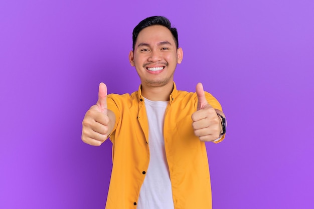 Un joven asiático sonriente y apuesto que muestra un gesto con el pulgar hacia arriba mirando una cámara aislada en un fondo morado