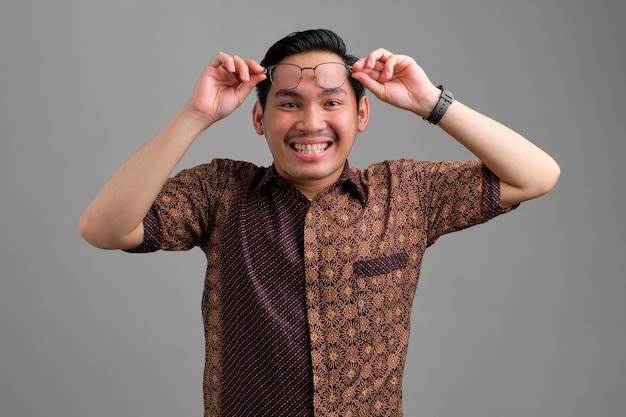 Un joven asiático sonriente con camisa de batik quitándose las gafas y mirando una cámara aislada de fondo gris