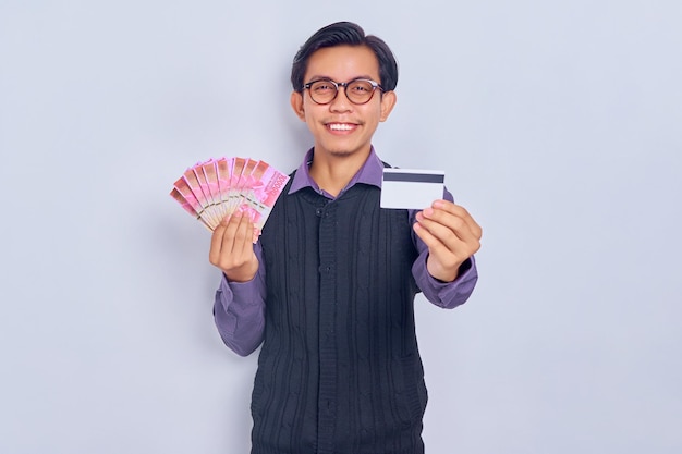 Un joven asiático sonriente con camisa de chaleco que muestra billetes de dinero en rupias y tarjeta de crédito aislado de fondo blanco