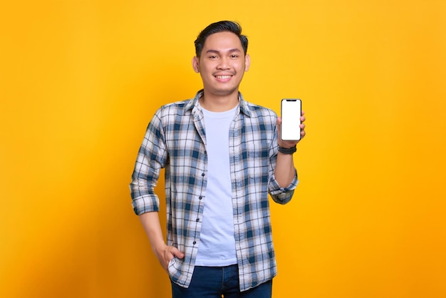 Un joven asiático sonriente con una camisa a cuadros que muestra una pantalla en blanco de un teléfono móvil recomendando una aplicación aislada de fondo amarillo