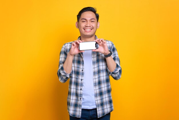 Un joven asiático sonriente con una camisa a cuadros que muestra una pantalla en blanco de un teléfono móvil recomendando una aplicación aislada de fondo amarillo