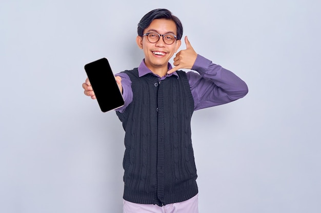 Un joven asiático sonriente con camisa informal y chaleco que muestra un teléfono móvil con pantalla en blanco y me hace un gesto aislado en el fondo blanco Concepto de estilo de vida de la gente