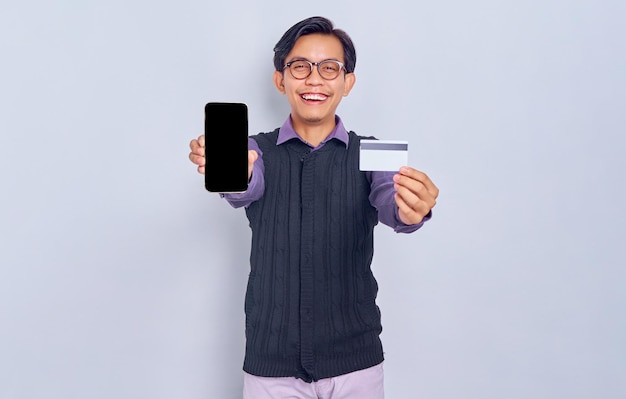Un joven asiático sonriente con camisa informal y chaleco que muestra un teléfono móvil con pantalla en blanco y sostiene una tarjeta de débito aislada en el fondo blanco Concepto de estilo de vida de la gente