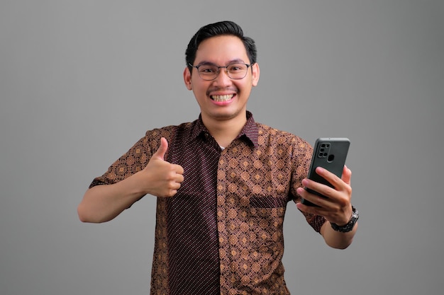 Un joven asiático sonriente con pantalones batik sosteniendo un smartphone y haciendo un gesto de aprobación aislado de fondo gris