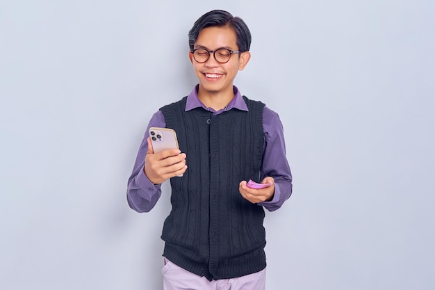 Un joven asiático sonriente con un pantalones casual usando un smartphone y sosteniendo billetes de dinero aislados de fondo blanco