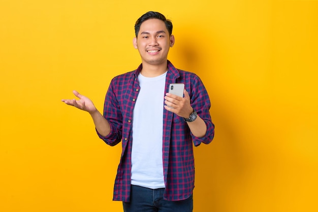 Un joven asiático sonriente con pantalones a cuadros usando un teléfono móvil leyendo noticias de una red social aislado de fondo amarillo