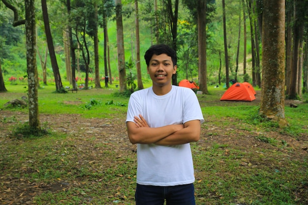 un joven asiático sonriente que usa una camiseta blanca con los brazos cruzados parado en el campamento es relajarse