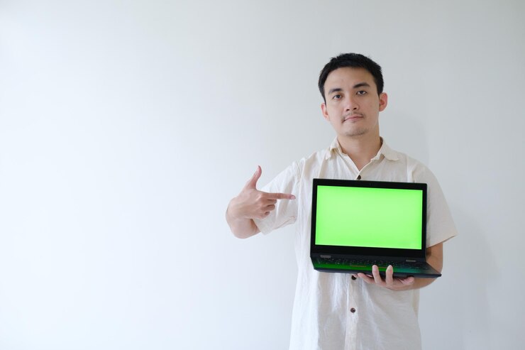 Un joven asiático sosteniendo una computadora portátil con una pantalla verde y mostrándosela a la cámara