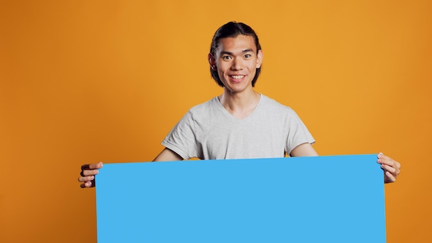 Foto joven asiático sosteniendo un tablero de cartón azul en la cámara, usando un espacio de copia aislado en una cartelera en el estudio. persona positiva alegre que muestra un icono de cartón con espacio en blanco, mensaje publicitario.