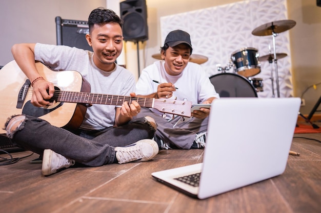 Joven asiático tocando la guitarra y su amigo viendo un video tutorial musical