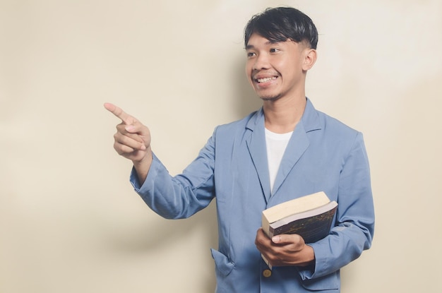 Joven asiático con traje universitario apuntando al espacio vacío mientras lleva un libro sobre un fondo aislado