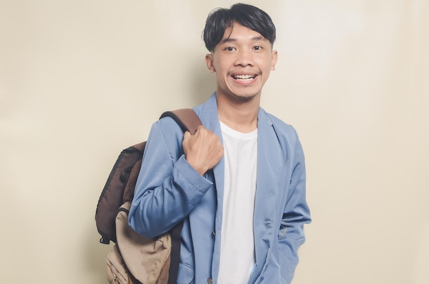 Joven asiático con traje universitario sonriendo mientras lleva una bolsa en un fondo aislado