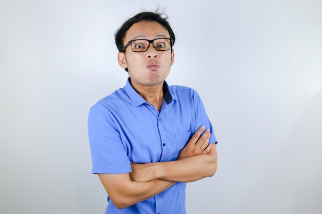 El joven asiático usa una camisa azul con una cara graciosa y enojada con los brazos cruzados, mira la cámara aislada sobre el fondo blanco