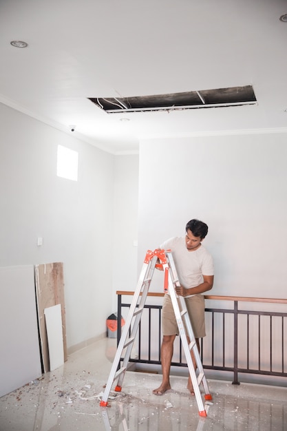 Joven asiático usando una escalera para reparar el techo roto en casa
