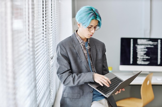 Joven asiático usando una laptop sentada junto a la ventana en la oficina