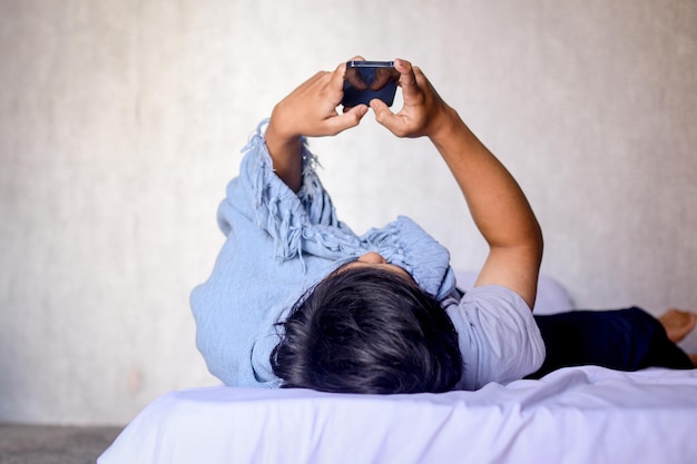 Joven asiático usando un mensaje de escritura telefónica para chatear en la red social mientras está acostado en la cama en b