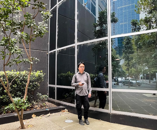 Foto joven asiático usando un teléfono inteligente mientras está de pie frente a una fachada de vidrio reflectante en la ciudad