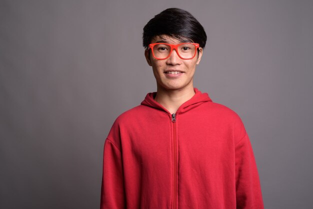 Foto joven asiático vistiendo chaqueta roja con anteojos contra gris