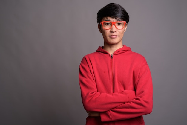 Joven asiático vistiendo chaqueta roja con anteojos contra gris