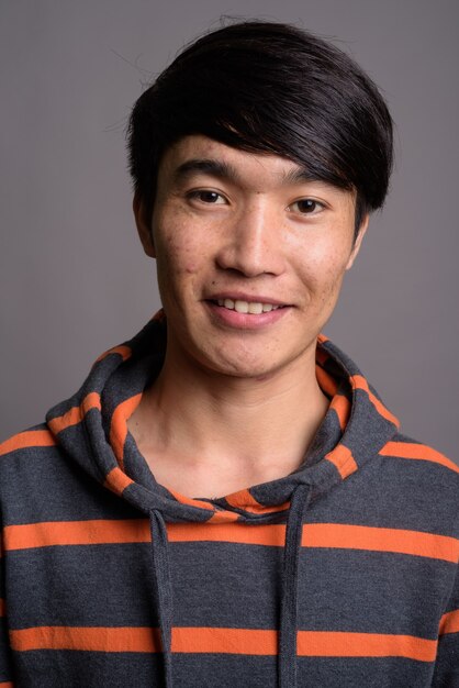Joven asiático vistiendo sudadera con capucha a rayas contra la pared gris