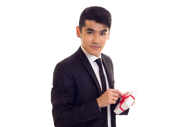 Un joven de aspecto elegante con el pelo negro con camisa blanca y traje negro con corbata sosteniendo un regalo de fondo blanco