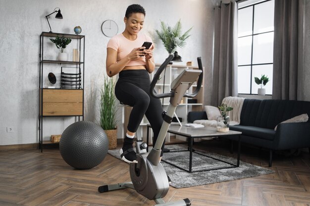 Joven atleta africana vistiendo ropa deportiva y usando bicicleta estática usando un teléfono inteligente